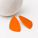 Bright orange geometric stud earrings with a matte finish, displayed on a clean white surface next to a white marble sphere.