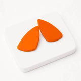 A pair of bright orange teardrop-shaped statement earrings displayed on a white square coaster against a white background.