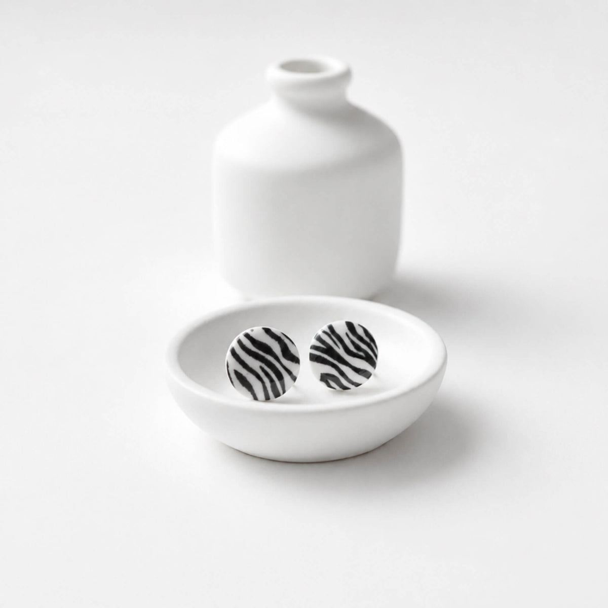 A pair of round stud earrings with a black and white zebra stripe pattern, displayed in a small white ceramic dish with a matching white ceramic vase in the background on a white surface.