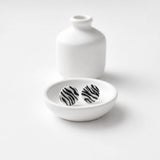 A pair of round stud earrings with a black and white zebra stripe pattern, displayed in a small white ceramic dish with a matching white ceramic vase in the background on a white surface.